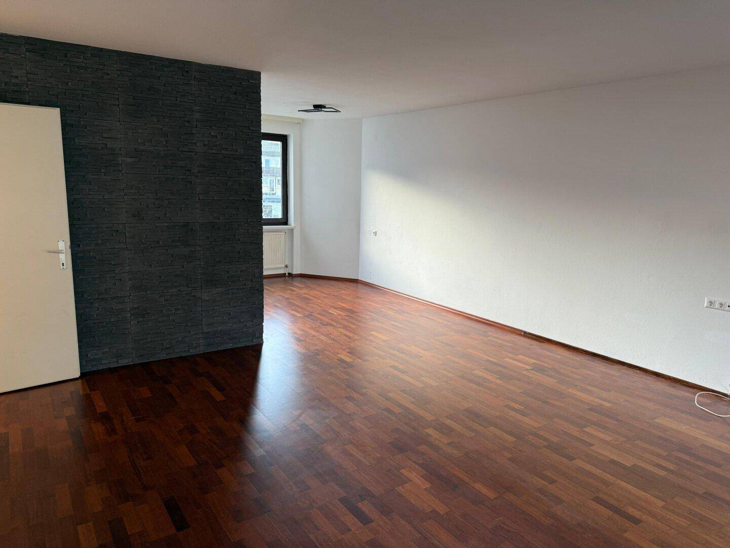 Geräumiges, leeres Wohnzimmer mit dunklem Laminatboden und einer modernen Akzentwand.