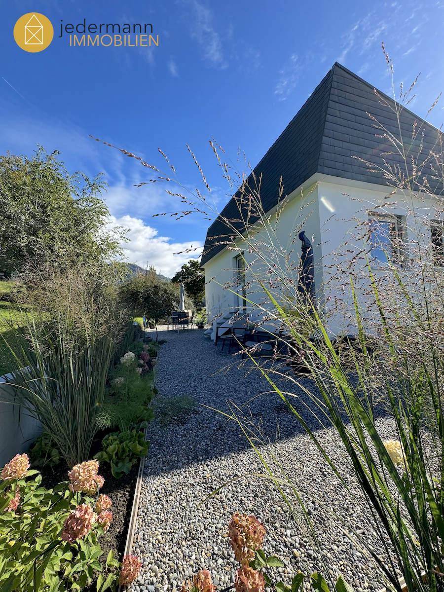Seitenansicht eines Hauses mit Kiesweg, Bepflanzung und Sitzgelegenheit im Hintergrund.