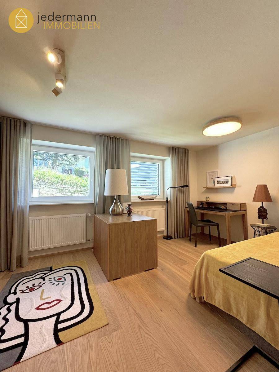 Schlafzimmer mit Holzboden, Schreibtisch und zwei Fenstern für viel Tageslicht.