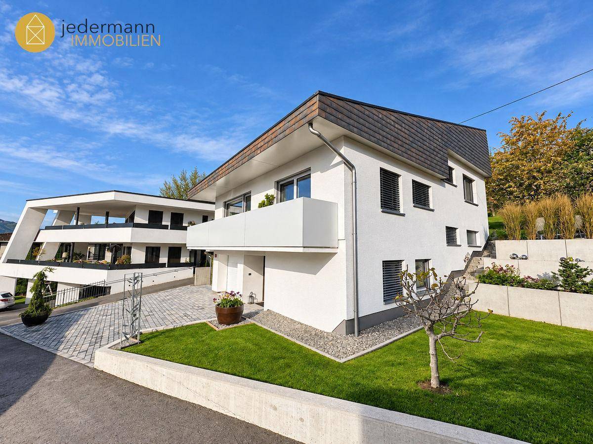 Modernes Einfamilienhaus mit weißer Fassade, Balkon und gepflegtem Vorgarten in grüner Umgebung.