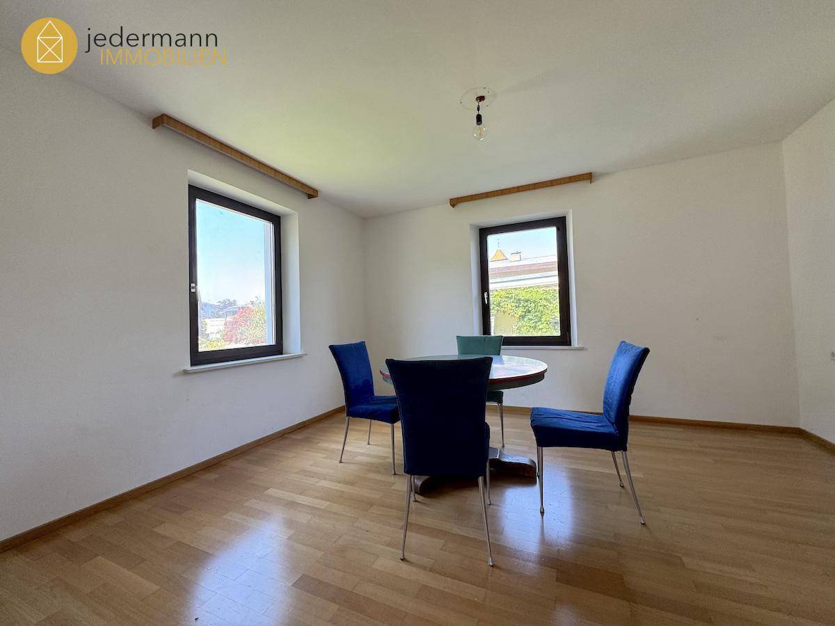 Esszimmer mit Holzboden, zwei Fenstern und einem runden Esstisch mit blauen Stühlen.