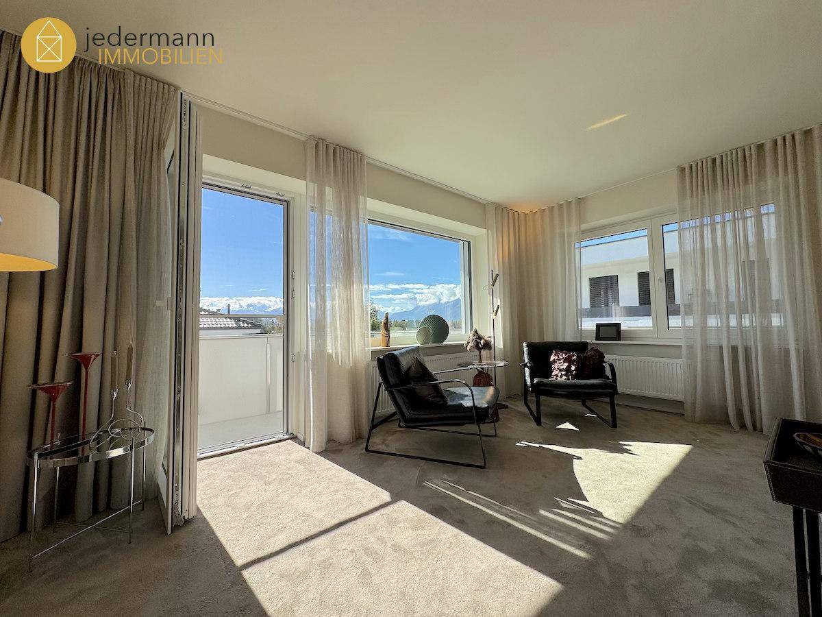 Helles Wohnzimmer mit großen Fenstern, Teppichboden und direktem Zugang zu einem Balkon mit Bergblick.