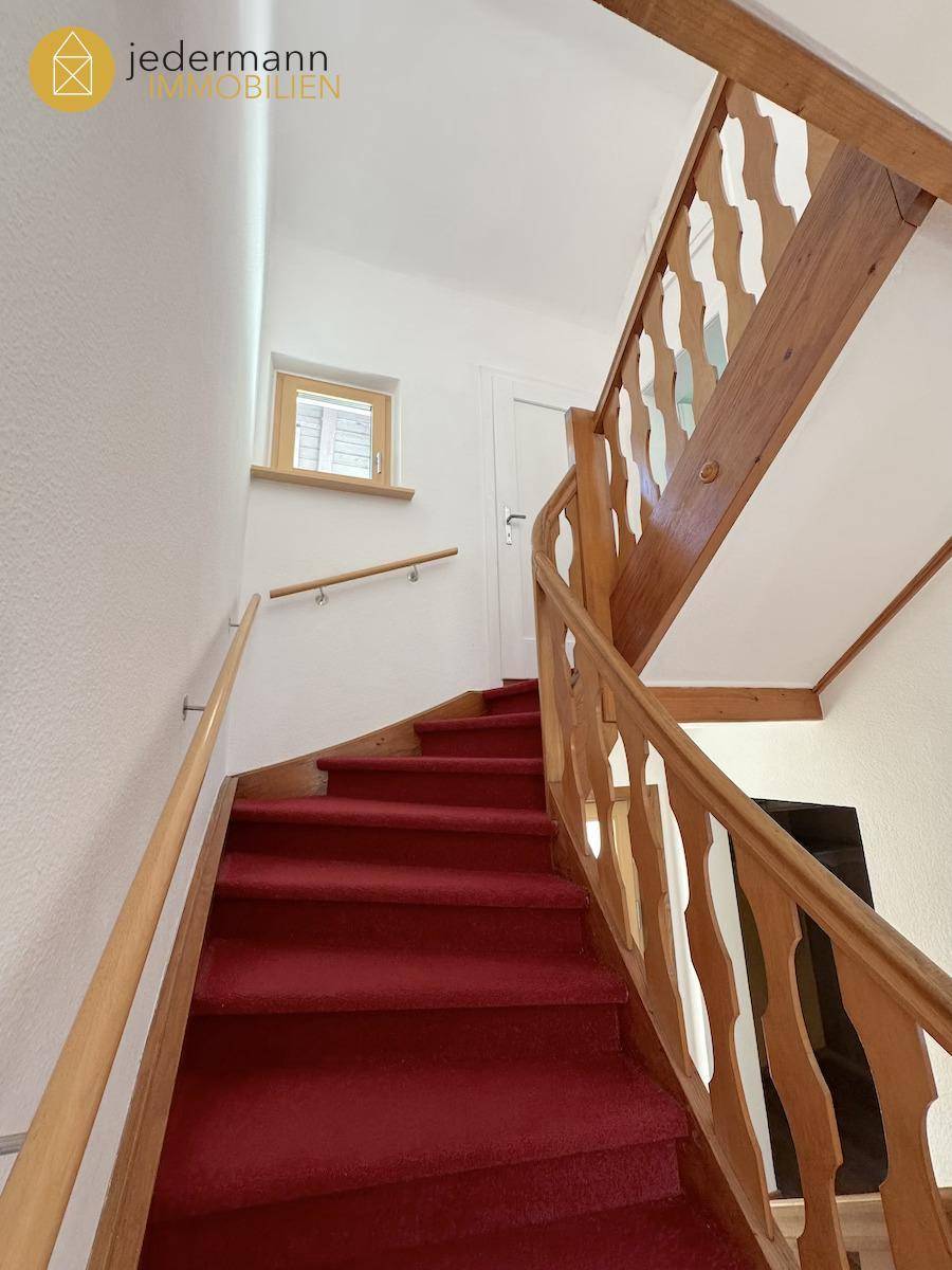 Holztreppe mit rotem Teppich und geschnitztem Geländer, die in die obere Etage führt.