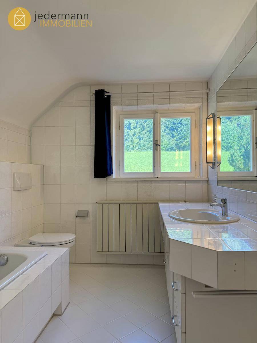 Gefliestes Badezimmer mit Badewanne, Waschbecken und Fenster mit Blick ins Grüne.
