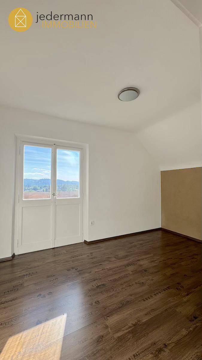 Helles Zimmer mit dunklem Laminatboden und Fenstertür mit weitem Ausblick.
