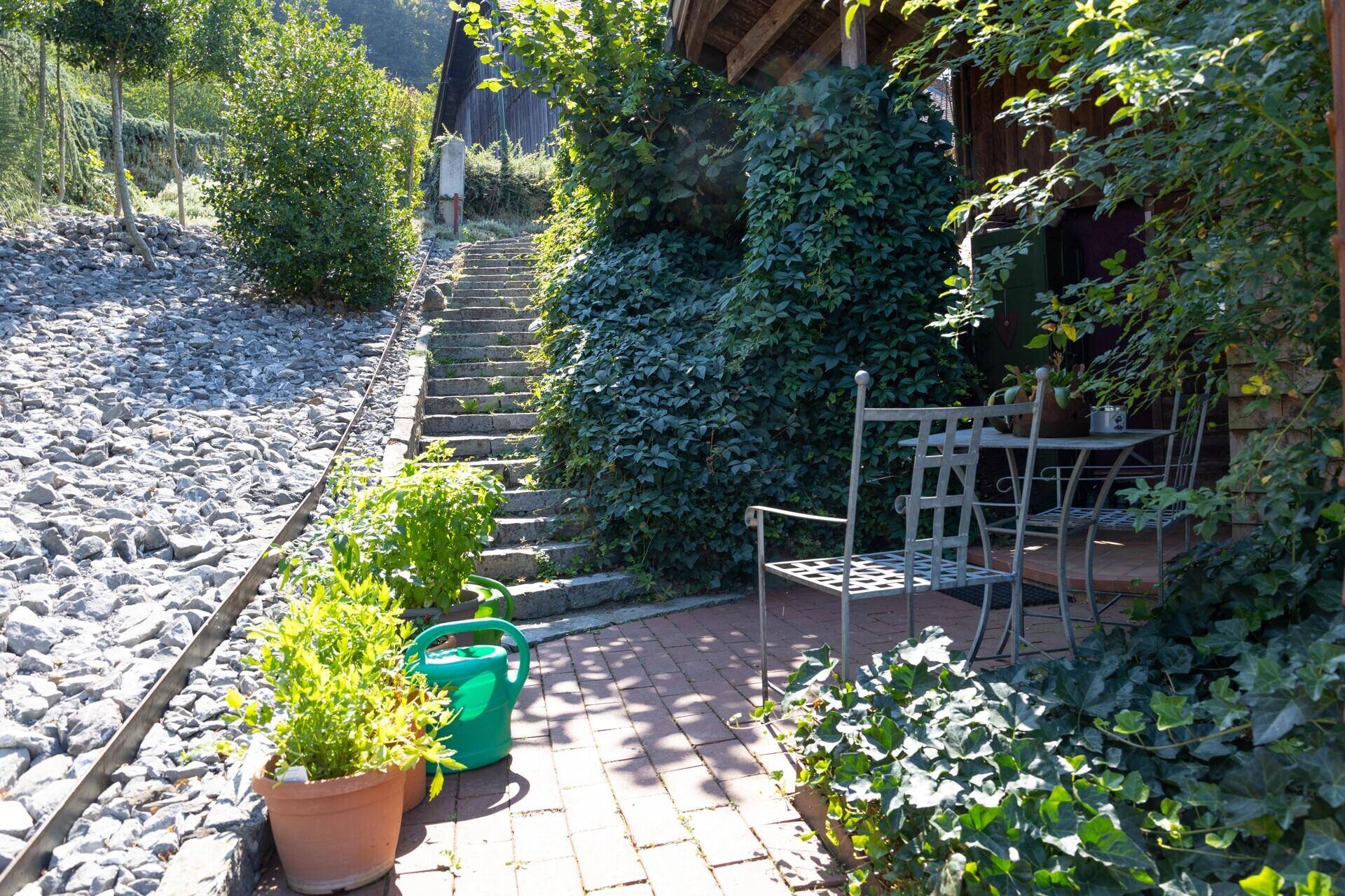 Gemütliche Terrasse mit Gartenmöbeln und üppiger Bepflanzung, umgeben von Natur und einem steinigen Weg.