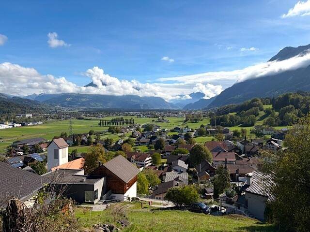 Weitläufiger Blick über ein idyllisches Dorf, umgeben von grünen Feldern und majestätischen Bergen.