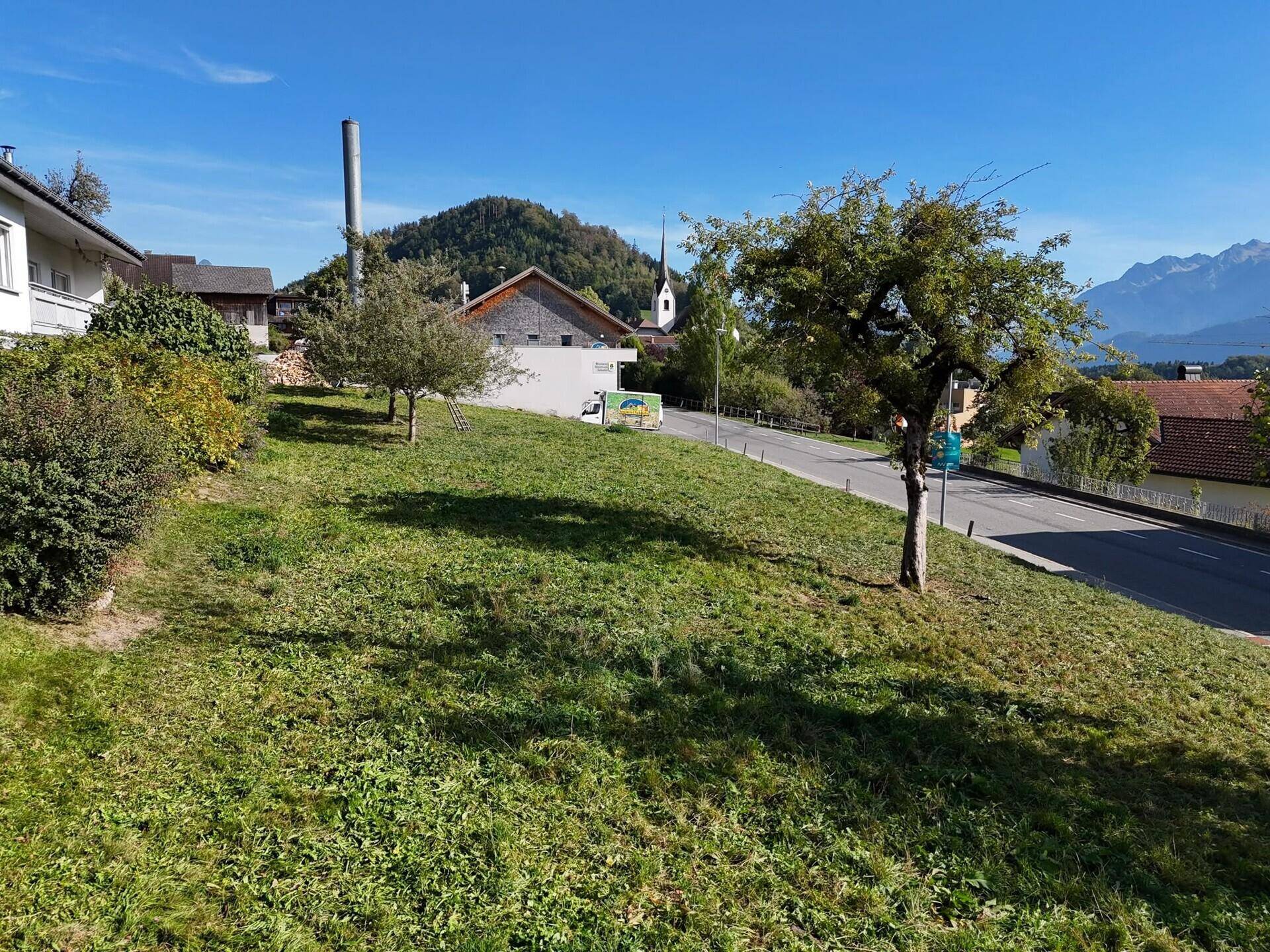 Grüne Parzelle mit Bäumen, angrenzend an eine Straße und mit Bergblick im Hintergrund.
