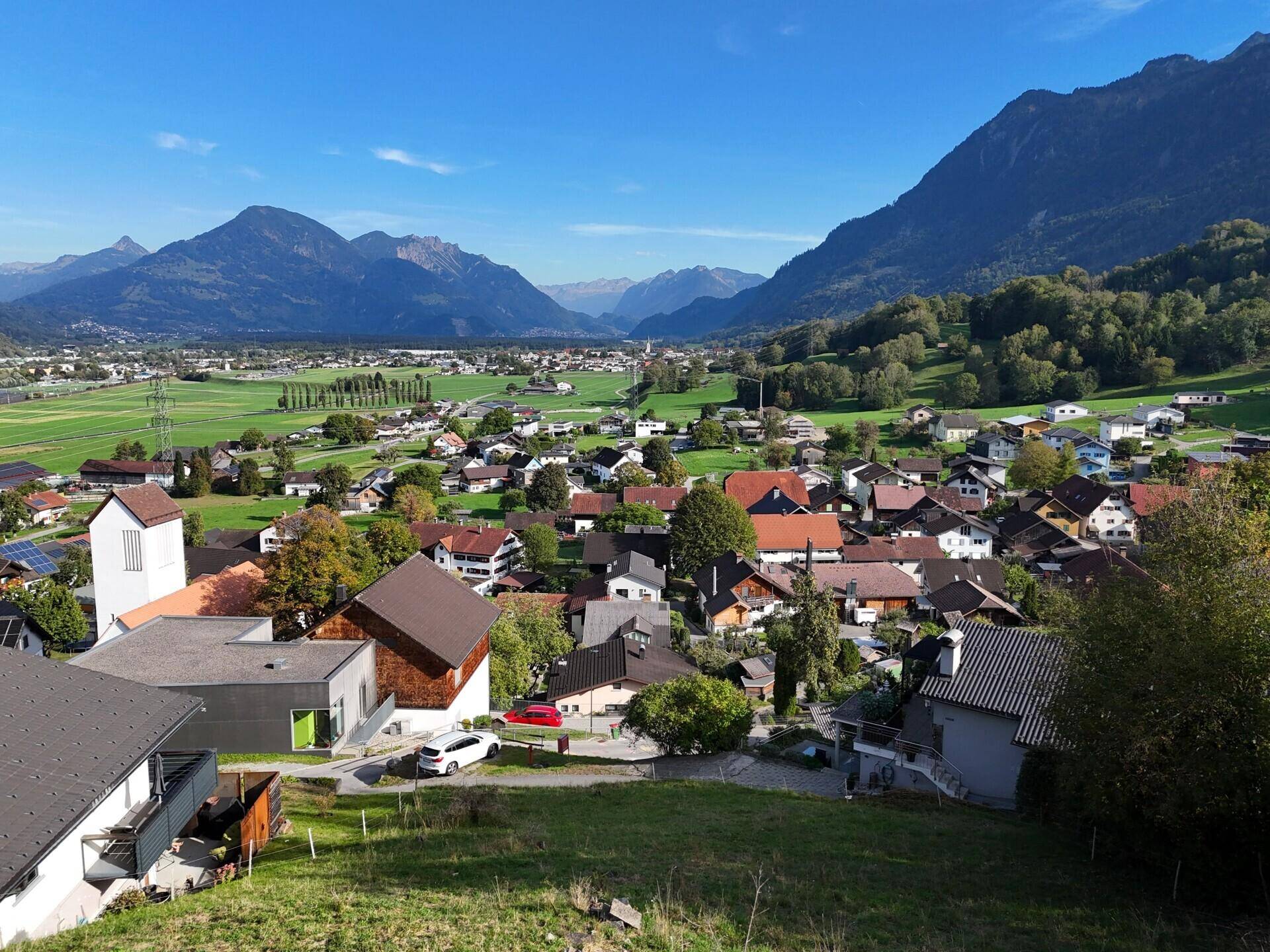 Sonniger Ausblick auf eine ländliche Siedlung mit Häusern, Feldern und einer beeindruckenden Bergkulisse.