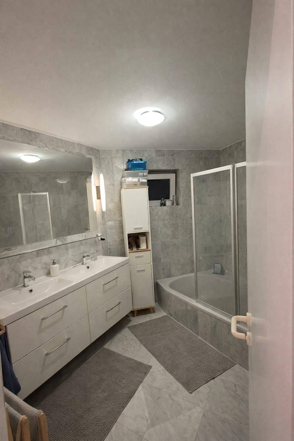 Modernes Badezimmer mit Doppelwaschbecken, Badewanne und grauen Fliesen, gut beleuchtet.