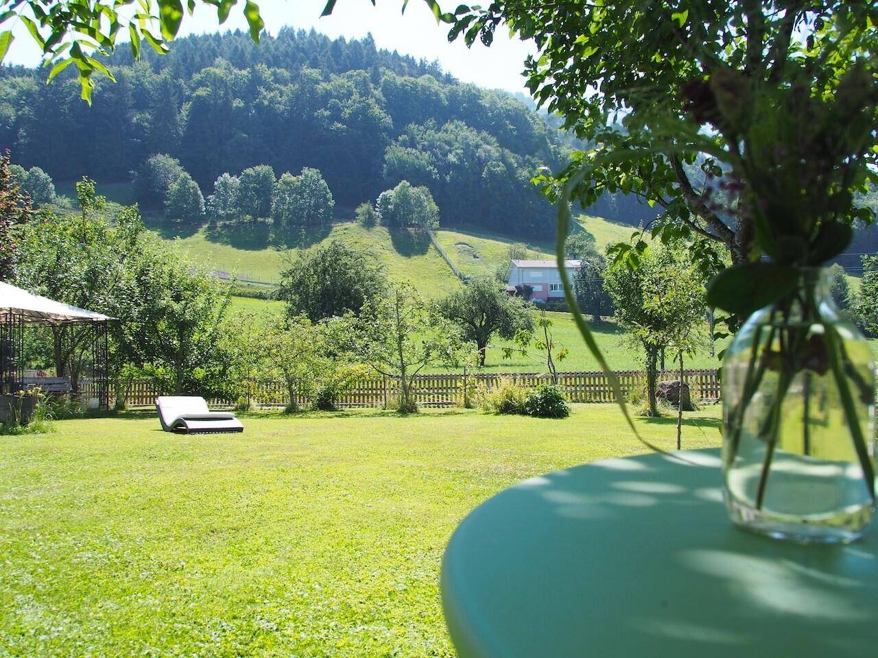 Idyllischer Garten mit Liegestuhl und weitem Blick auf grüne Hügel und bewaldete Landschaft.