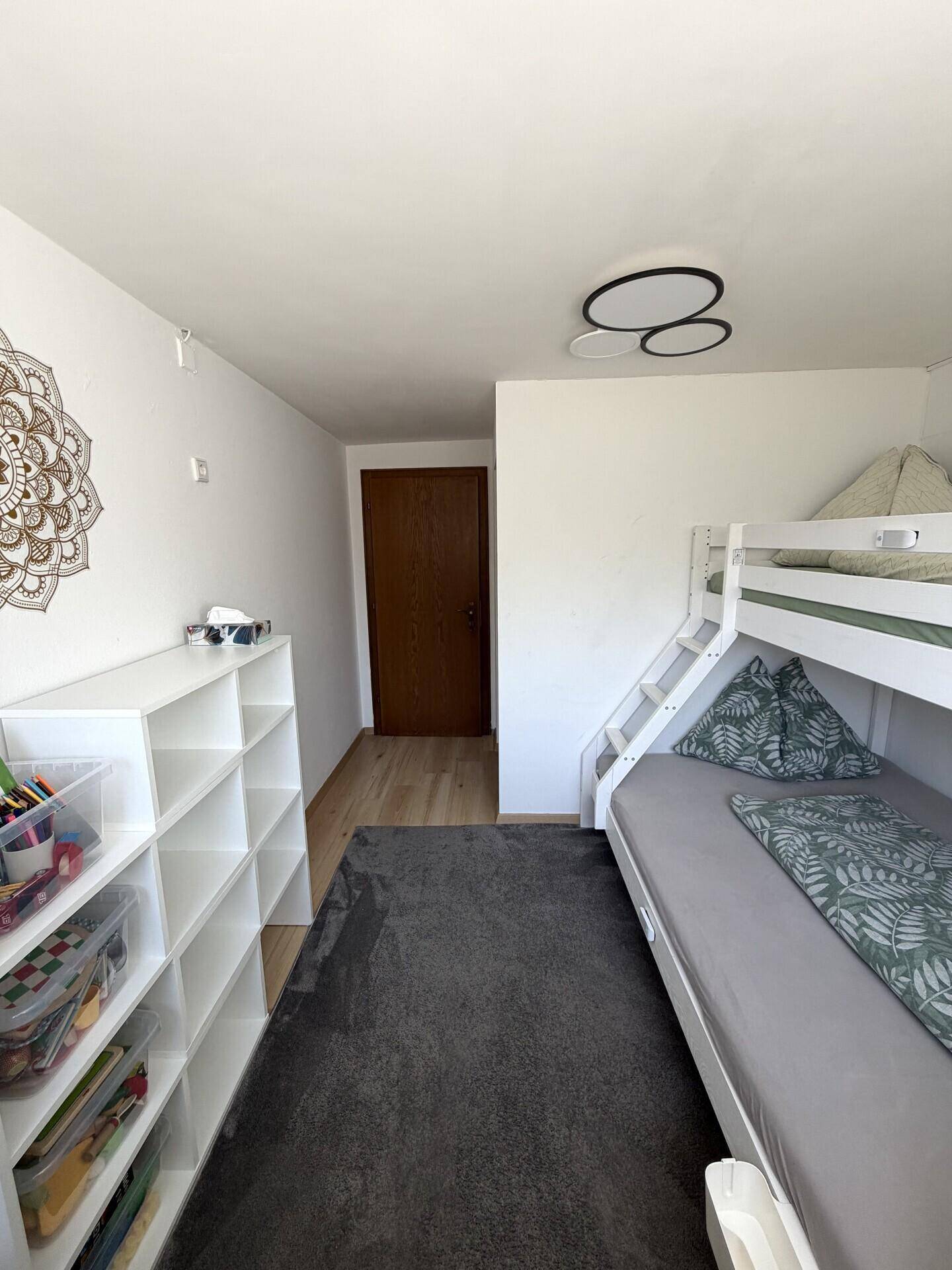 Helles Kinderzimmer mit Etagenbett, Regal und Teppichboden.