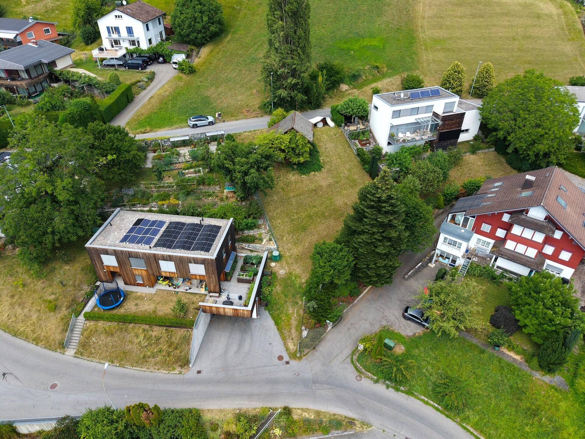 Luftaufnahme einer Wohnsiedlung mit mehreren Häusern, grünen Hügeln und einer Straße.