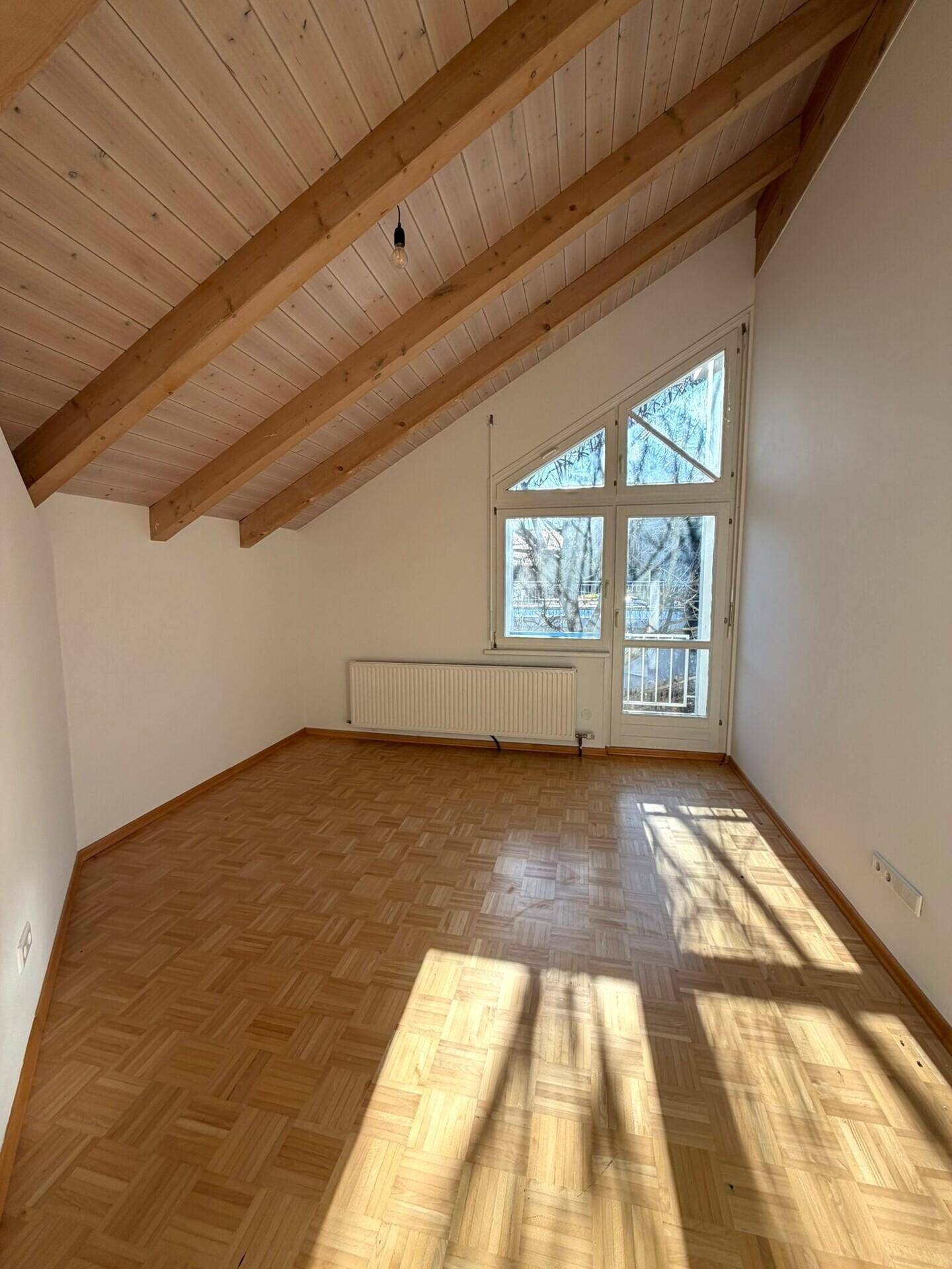 Helles Zimmer mit Parkettboden, schrägen Decken und großem Fenster, bietet viel Tageslicht.