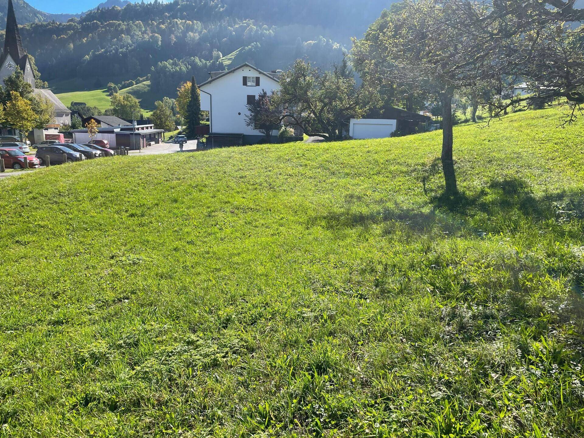 Sonniges grünes Grundstück mit Blick auf ein Dorf, eine Kirche und die majestätischen Berge in der Ferne.