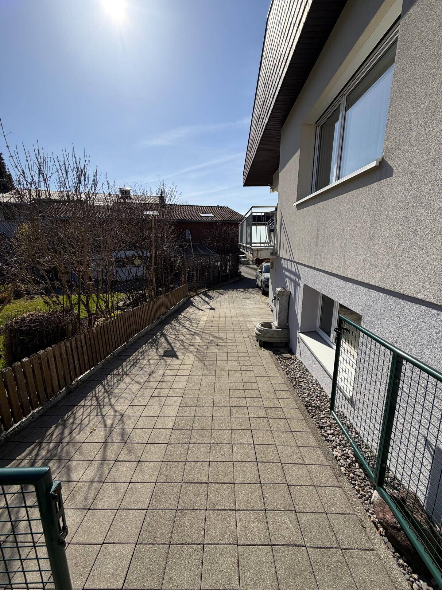 Gepflasterter Weg entlang der Hausseite mit einem kleinen Brunnen und Zaun.