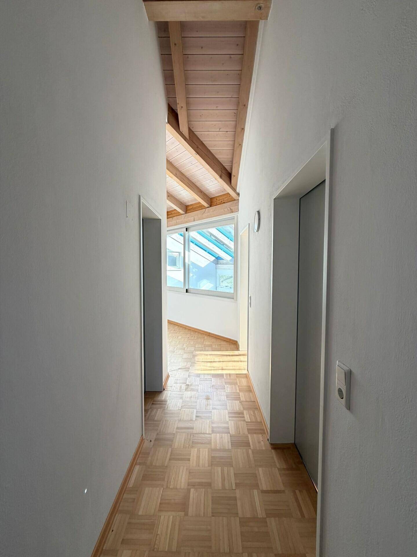Gangbereich mit Parkettboden und Holzdecke, der zu hellen Räumen mit großen Fenstern führt.