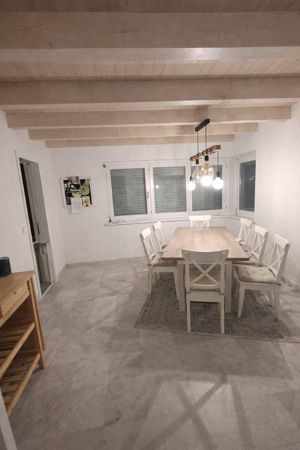 Heller Essbereich mit Holztisch, weißen Stühlen und sichtbaren Deckenbalken, auf Fliesenboden.