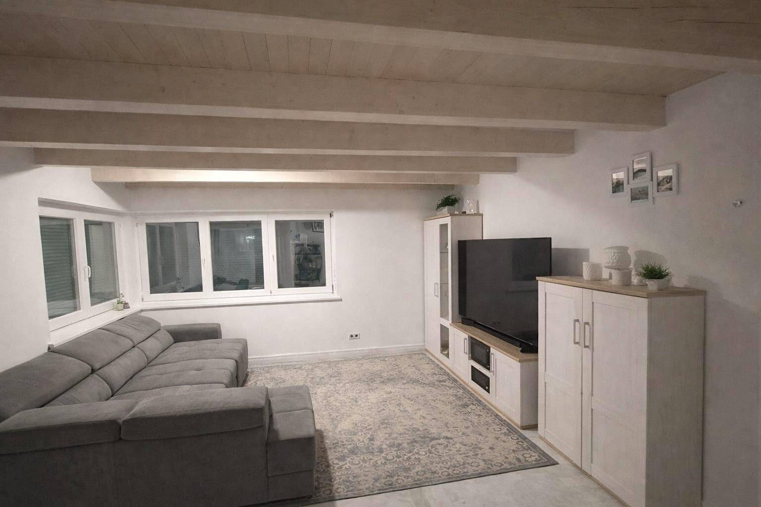 Gemütliches Wohnzimmer mit grauem Sofa, Teppich und sichtbaren Deckenbalken, gut beleuchtet.