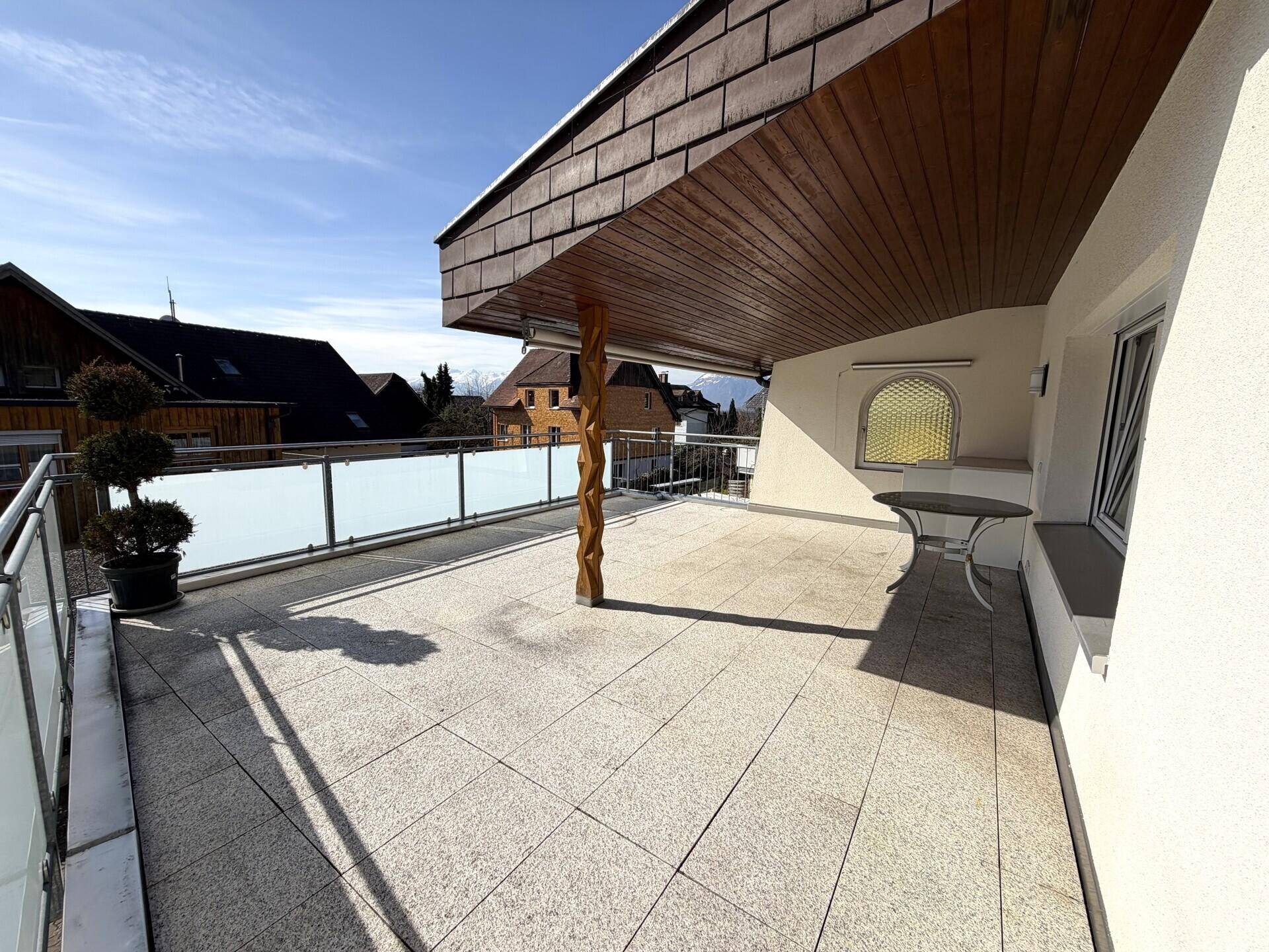 Weitläufige Terrasse mit Holzdach, Glasgeländer und Blick auf die Umgebung.