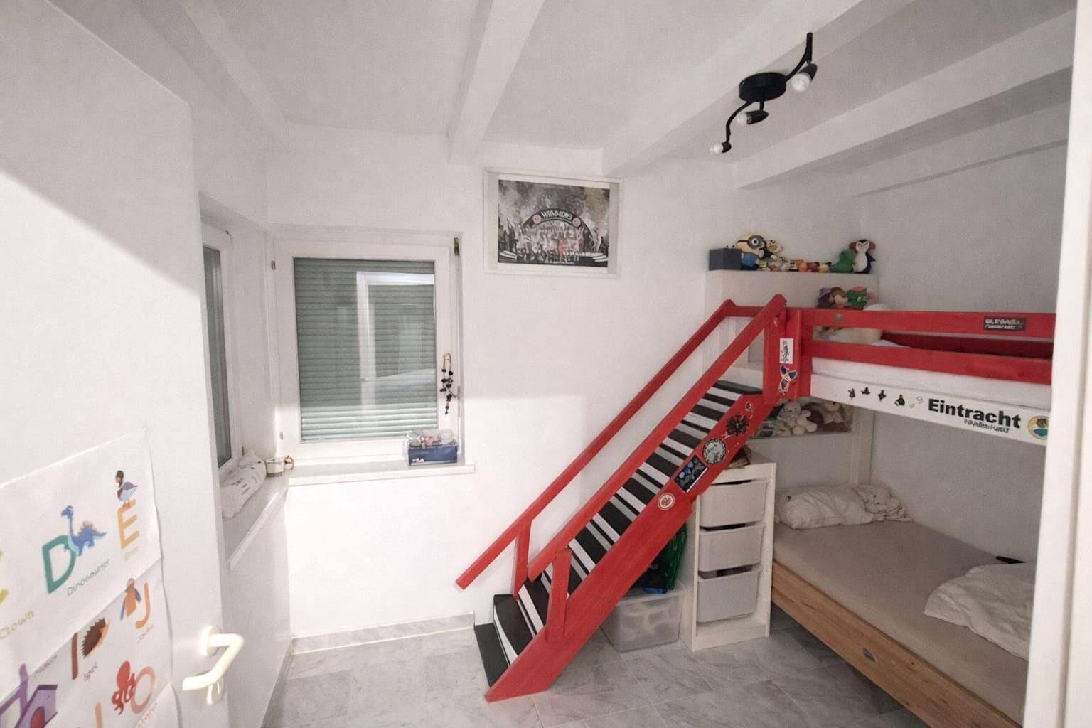 Kinderzimmer mit Etagenbett und roter Treppe, auf hellem Fliesenboden und mit Fenster.