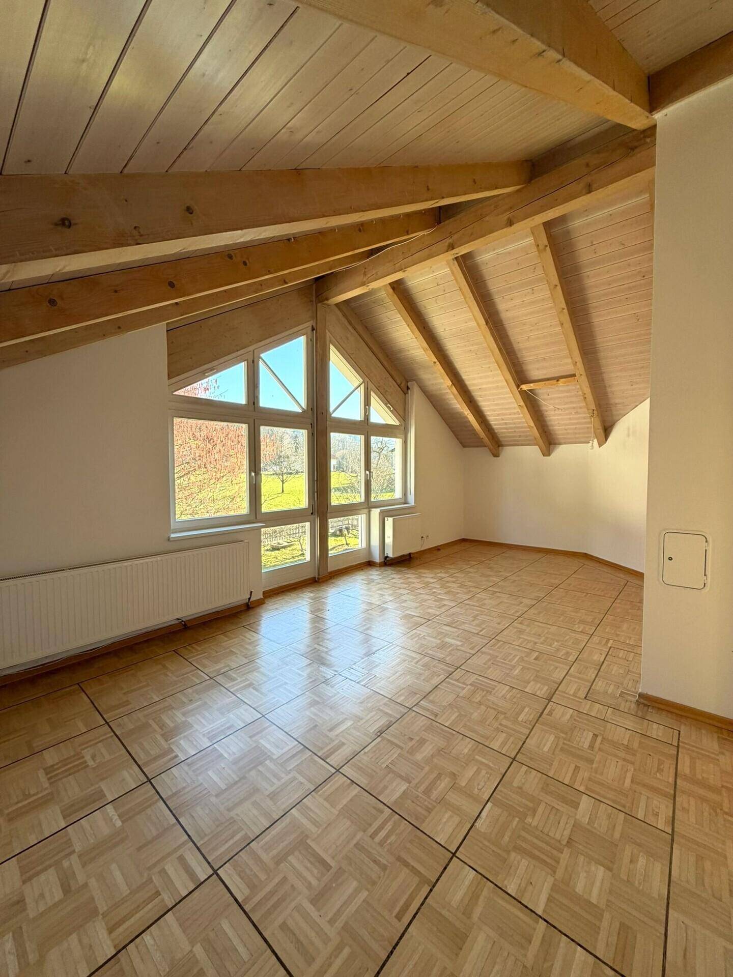 Geräumiger Wohnraum mit Parkettboden, hohen Fenstern und sichtbaren Holzdeckenbalken, lichtdurchflutet.