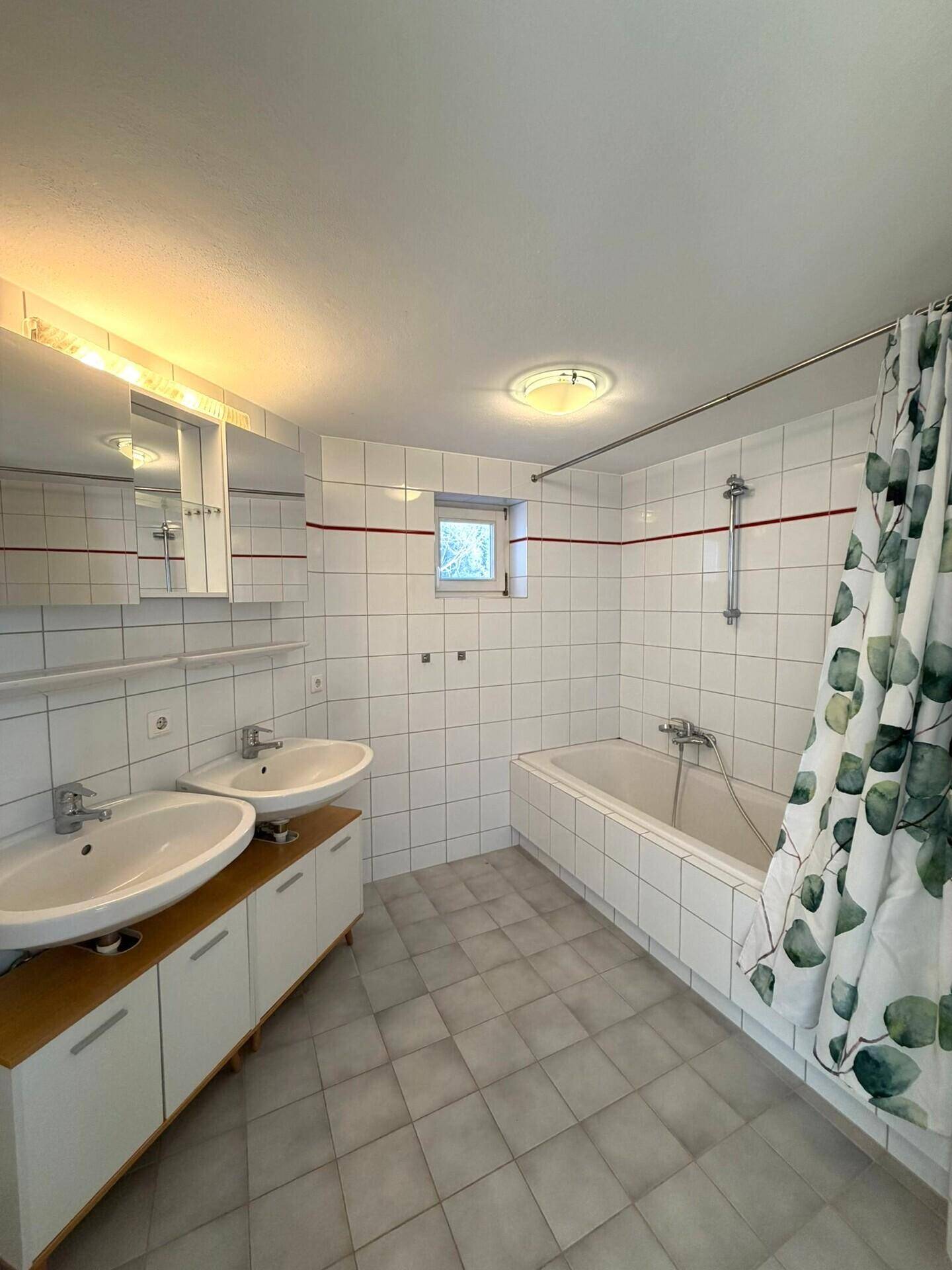 Helles Badezimmer mit Doppelwaschbecken, Badewanne und weißen Fliesen, ausgestattet mit Fenster.