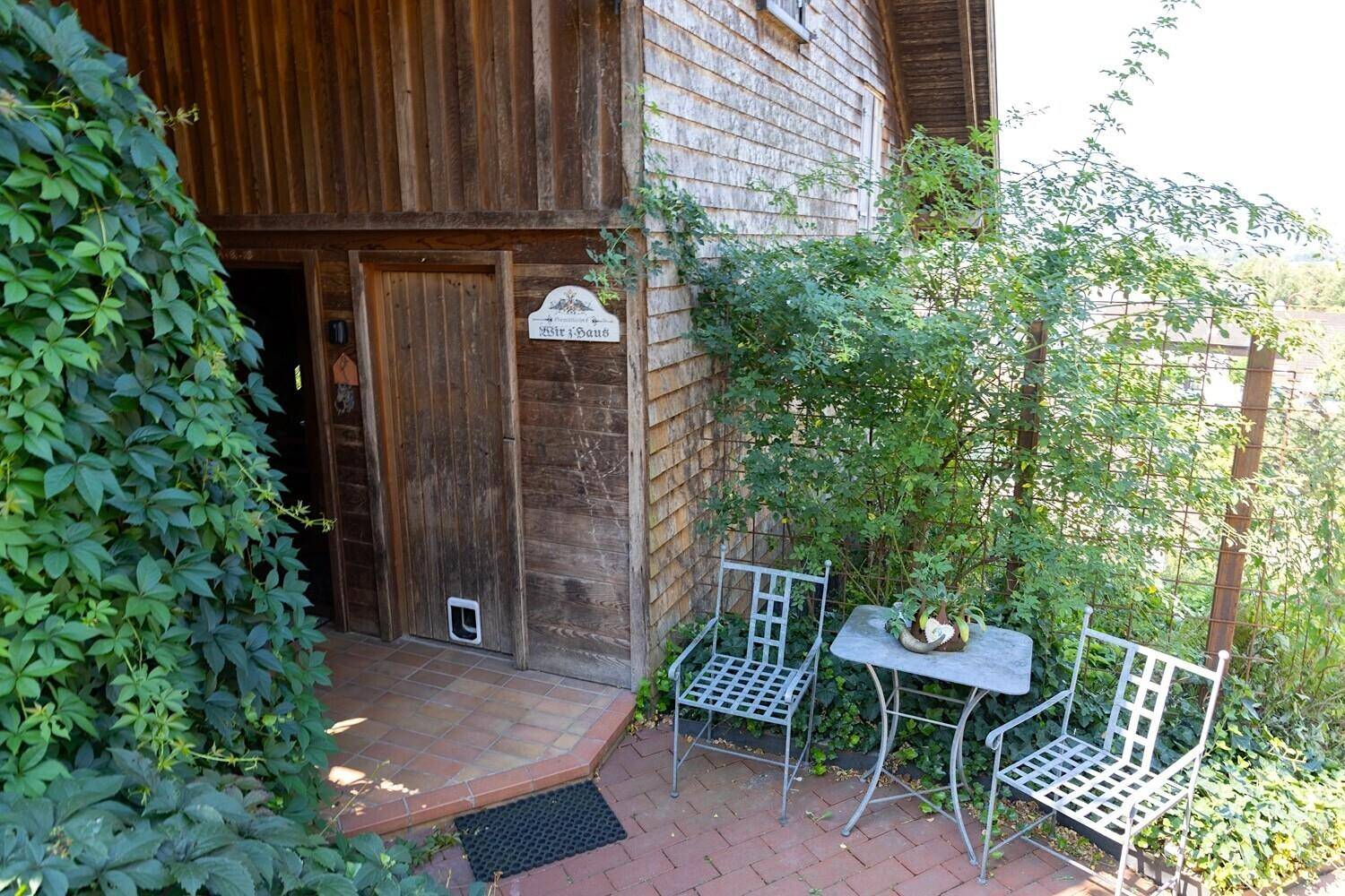 Eingangsbereich mit kleiner gepflasterter Terrasse und Sitzgelegenheiten, umgeben von Grün.