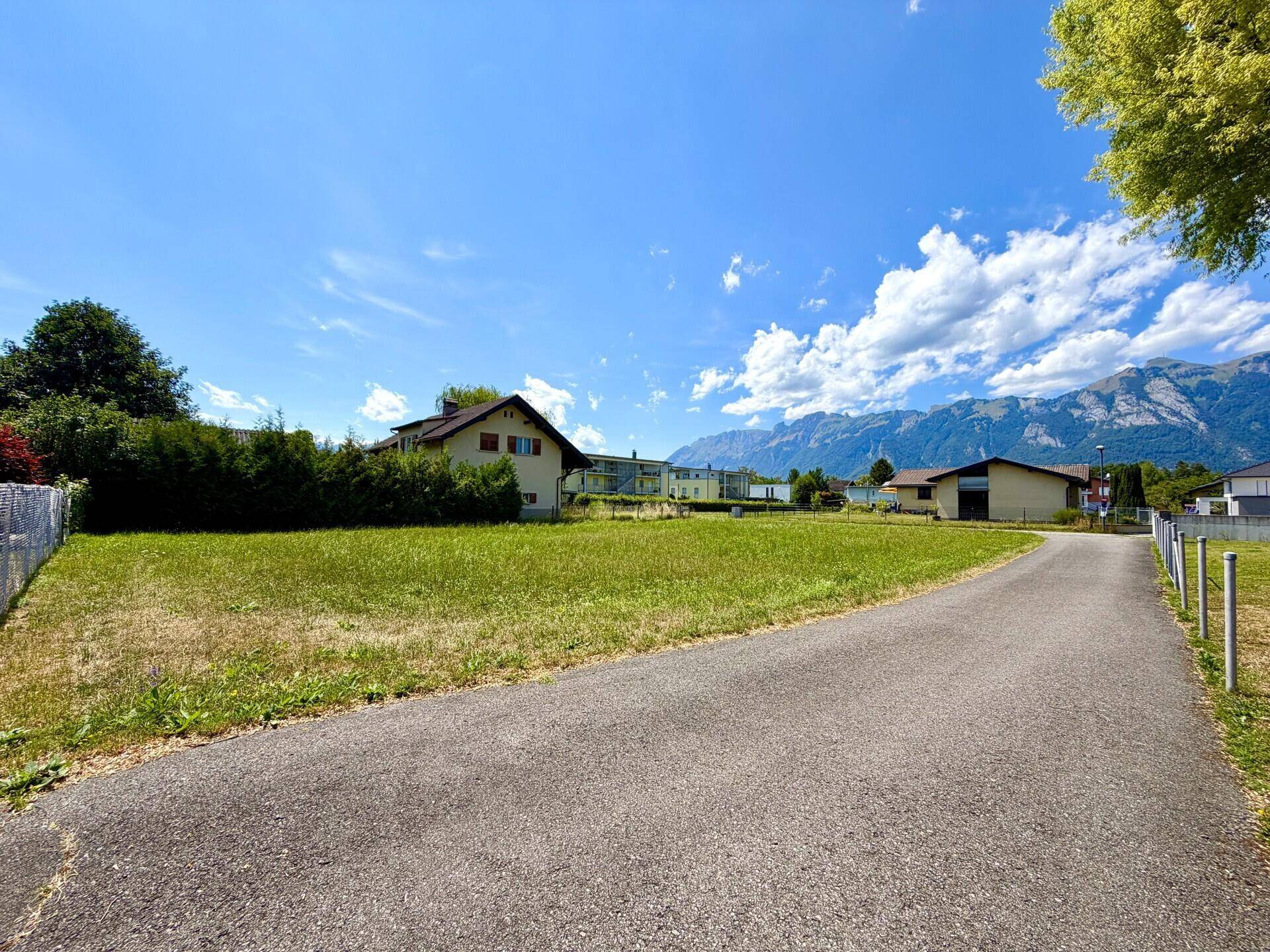 Offenes Grundstück mit grüner Wiese, umgeben von Wohngebäuden und mit weitem Blick auf die Berge.