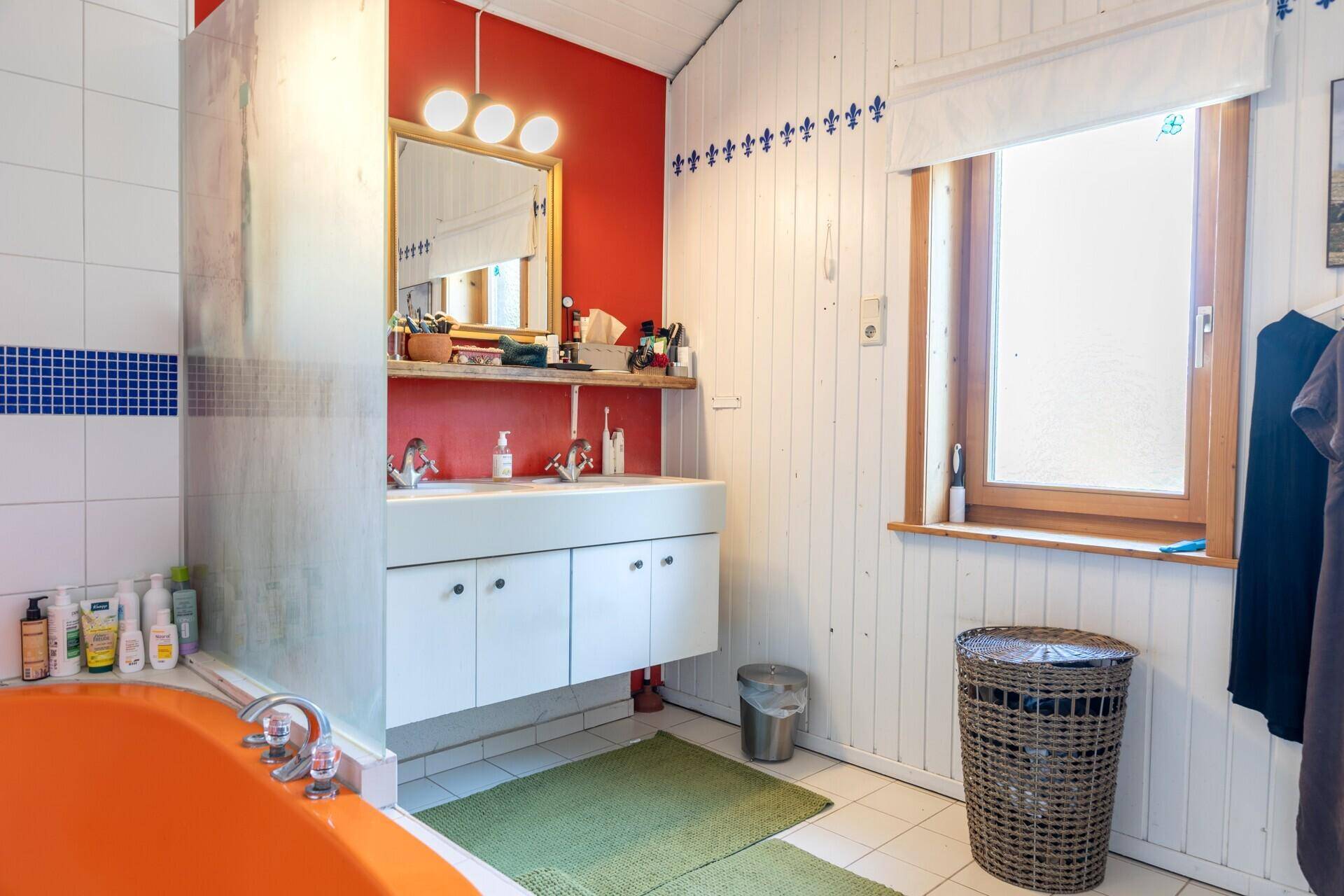 Badezimmer mit auffälliger orangefarbener Badewanne, Doppelwaschbecken und Fenster.