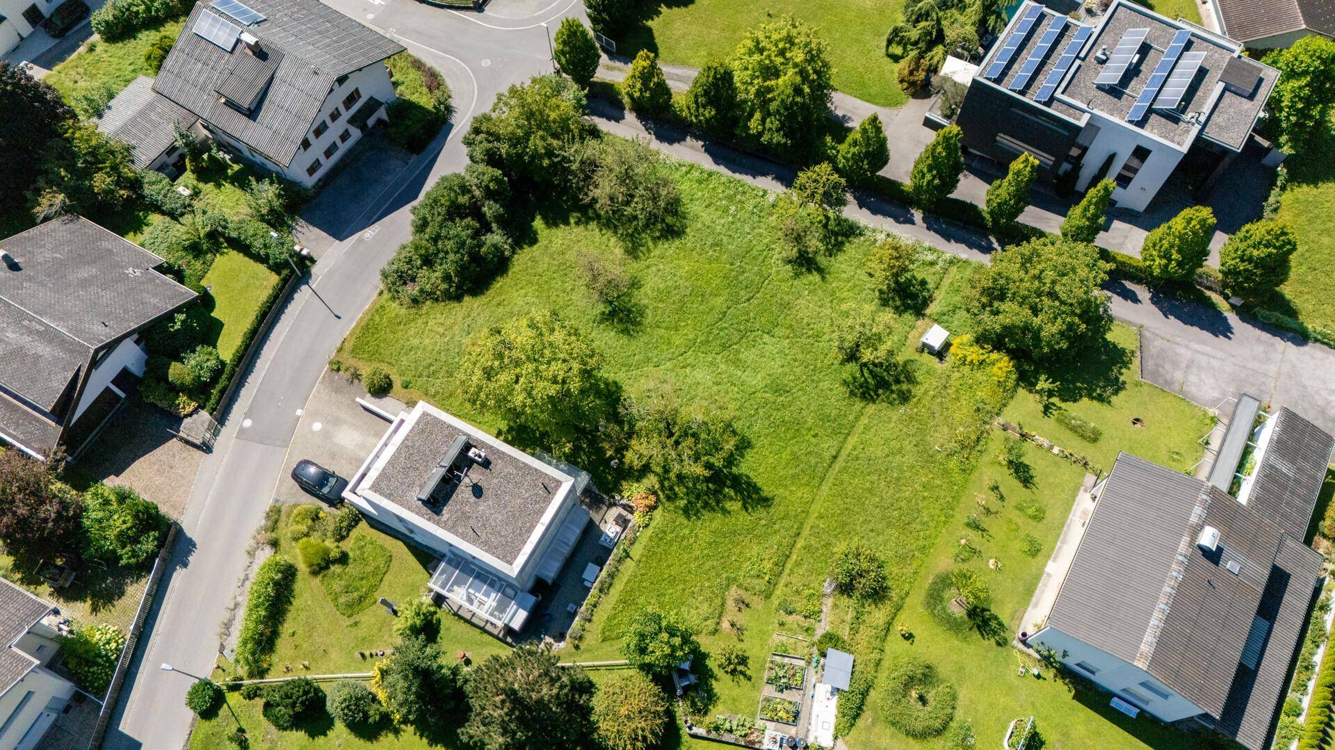 Luftaufnahme eines grünen Baugrundstücks mit umliegenden Häusern, einigen mit Photovoltaikanlagen auf dem Dach.
