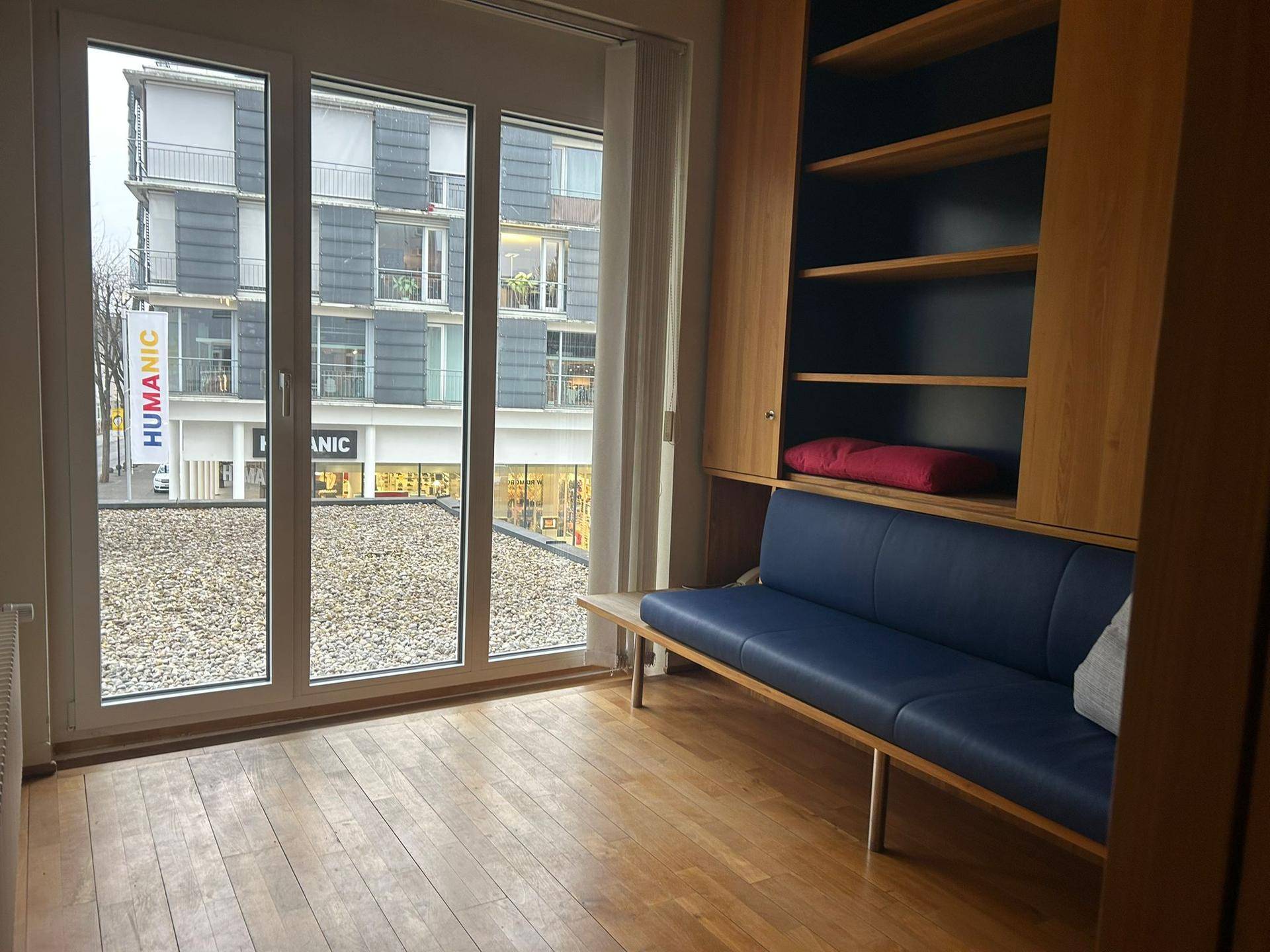 Heller Wohnraum mit Schlafsofa, Einbauregal und großem Fenster mit Blick auf die Stadt.