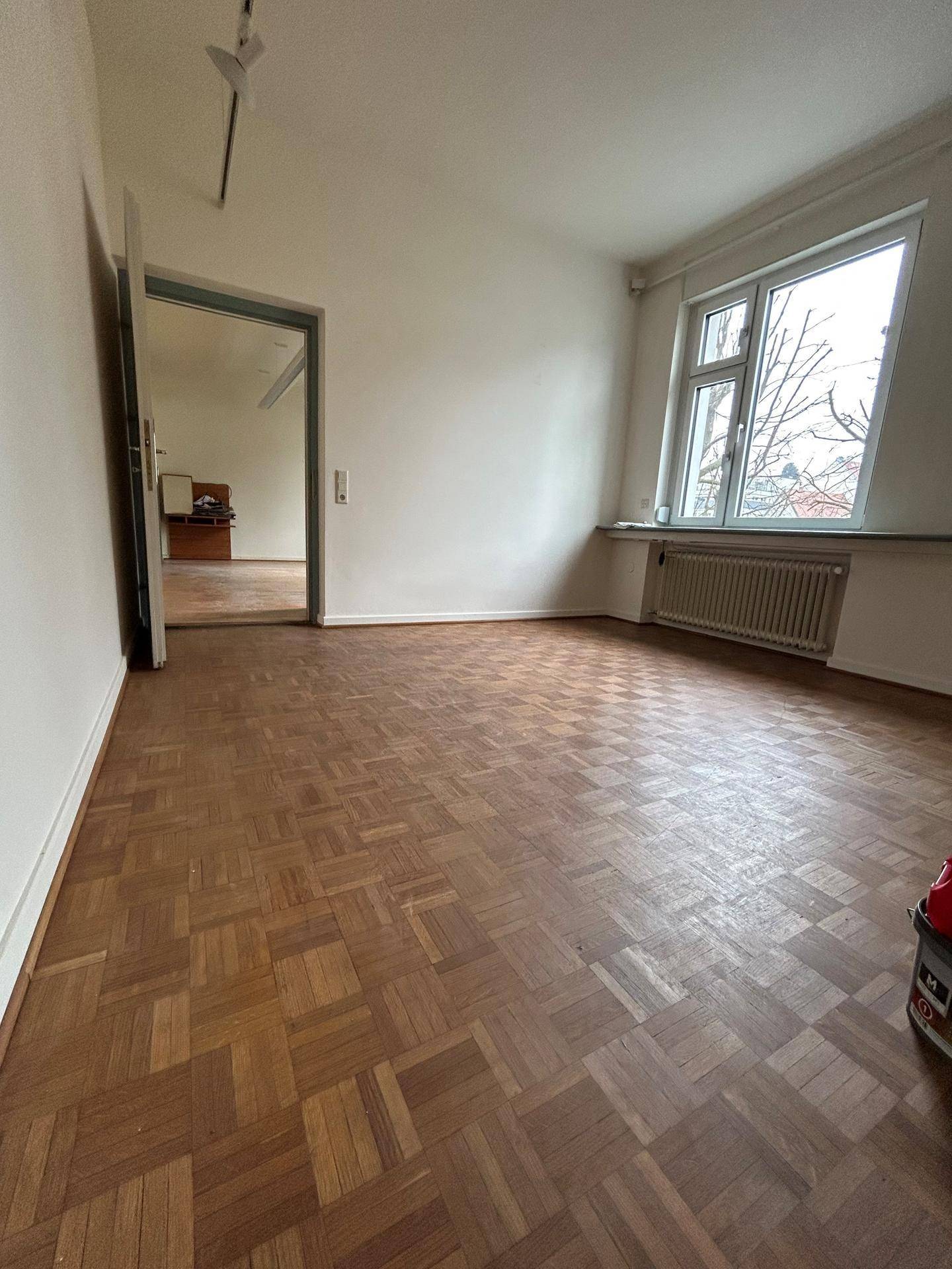 Geräumiger Wohnbereich mit Fischgrätparkett und einem Fenster, das Tageslicht hereinlässt.