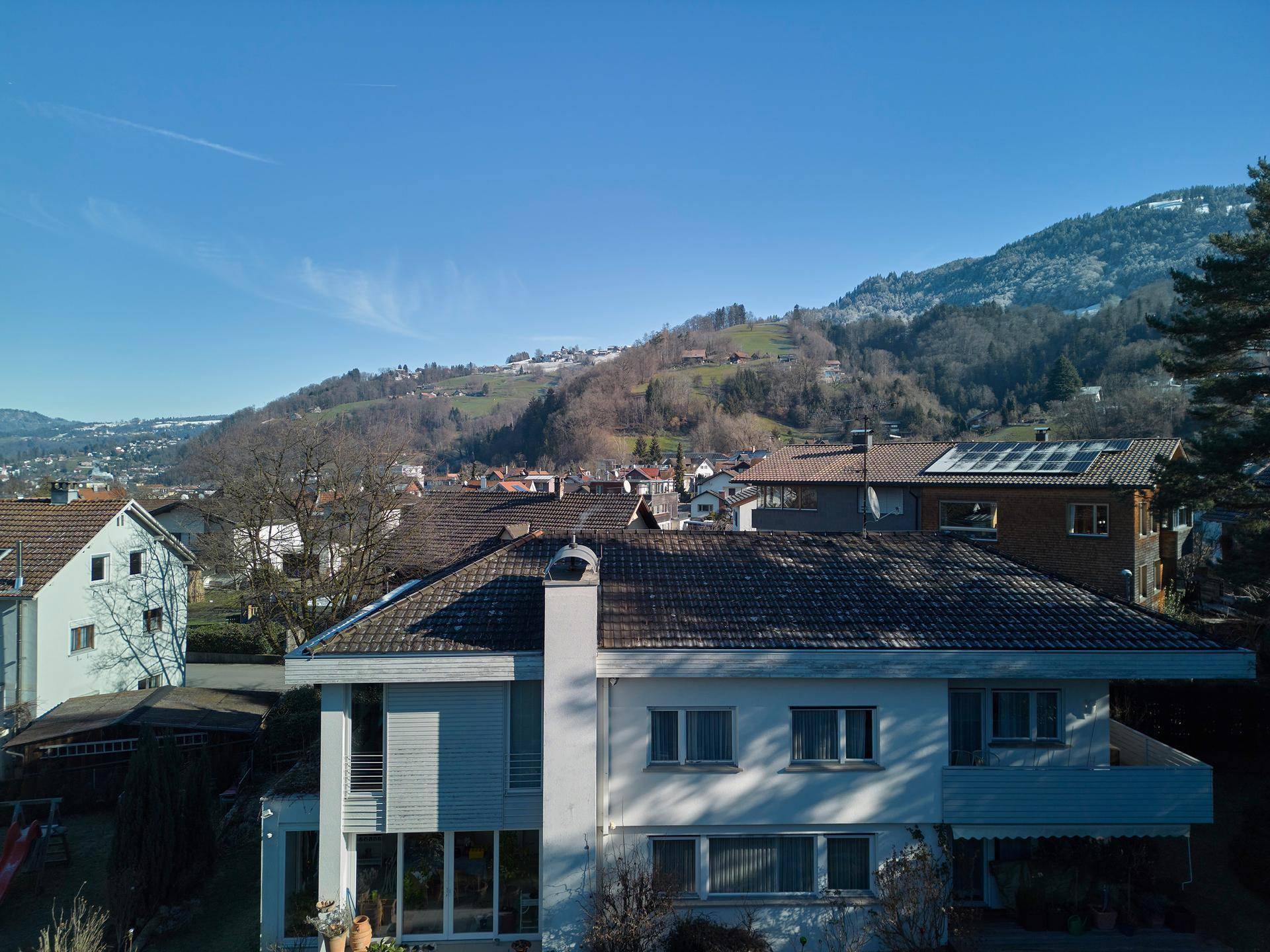 Luftaufnahme eines Hauses mit Ziegeldach und Schornstein, umgeben von grünen Hügeln und Wohnbebauung.