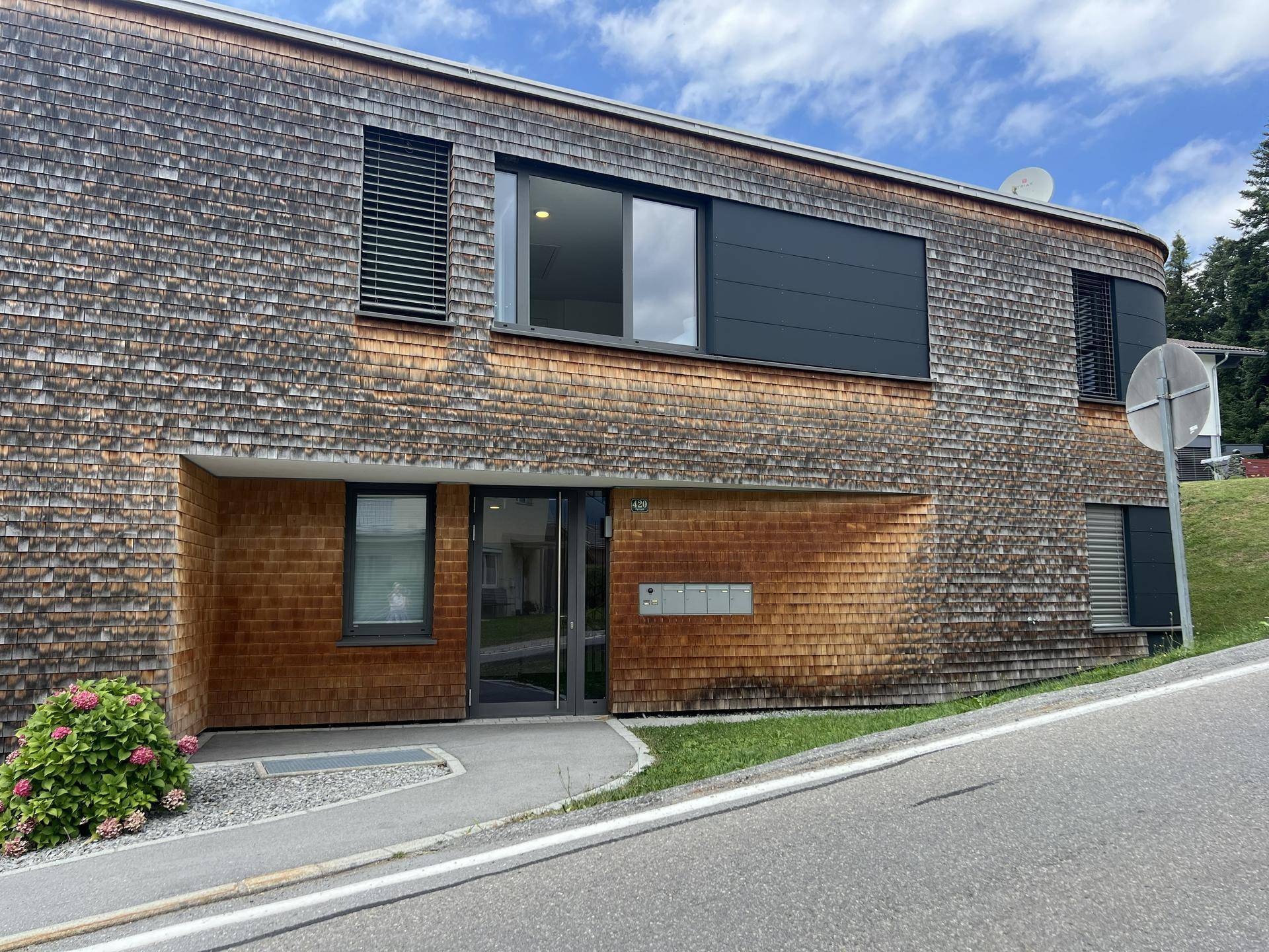 Eingangsbereich des modernen Gebäudes mit Holzschindelfassade und großen Fenstern.