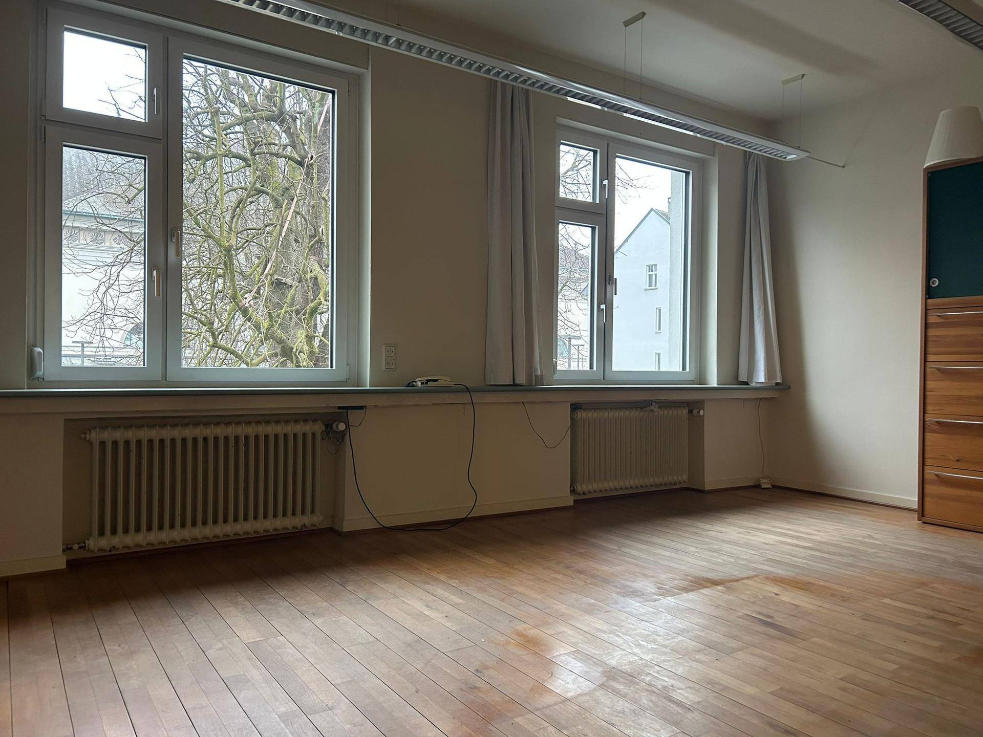 Heller, leerer Raum mit Holzboden, großen Fenstern und Blick auf Bäume und Gebäude.