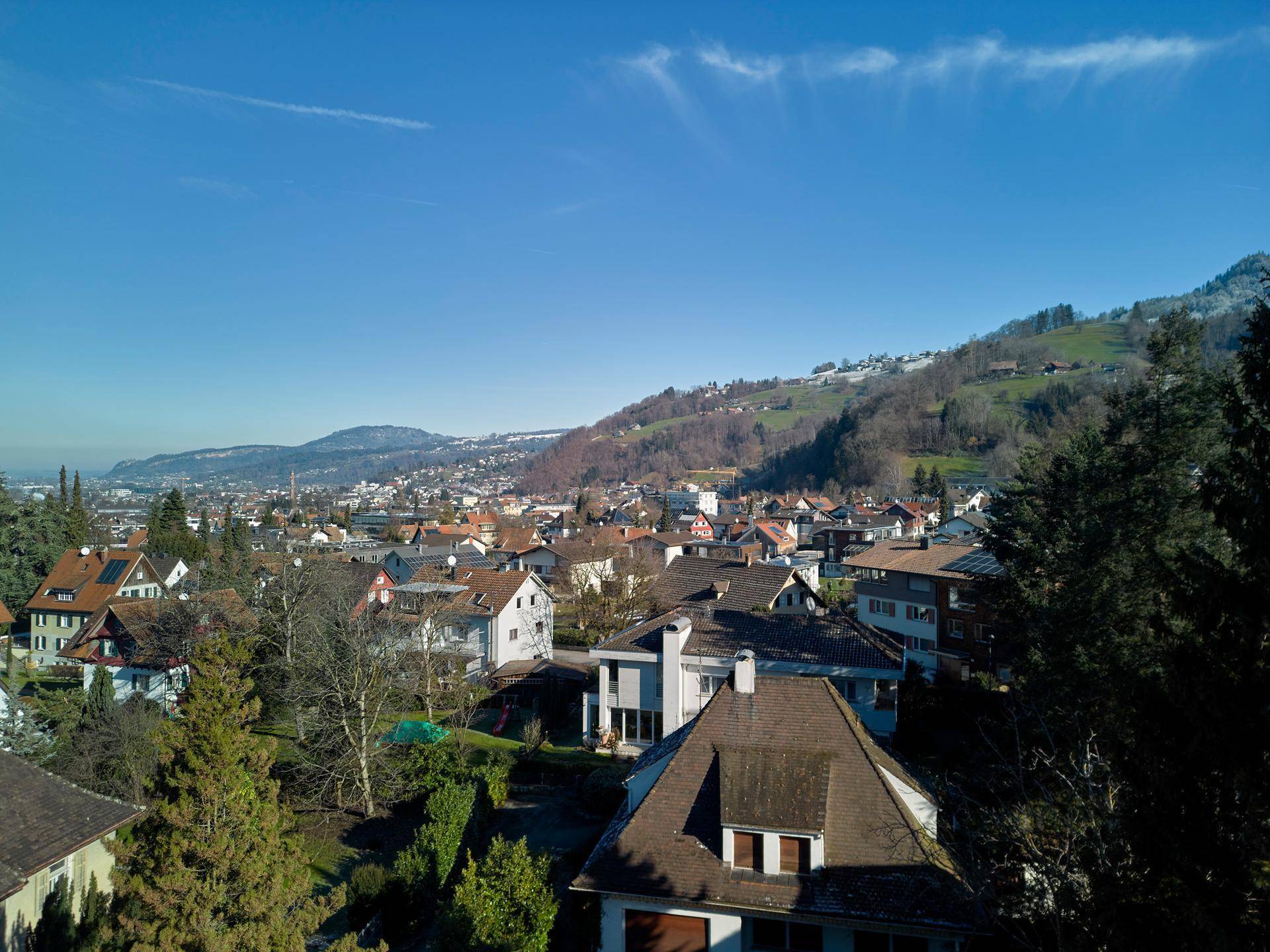 Luftaufnahme einer Wohnsiedlung mit Häusern und bewaldeten Hügeln im Hintergrund unter strahlend blauem Himmel.