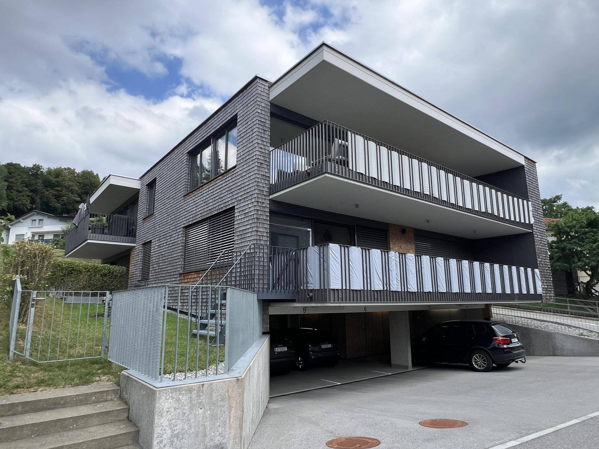 Modernes Mehrfamilienhaus mit Holzschindelfassade, Balkonen und Tiefgaragenzufahrt.