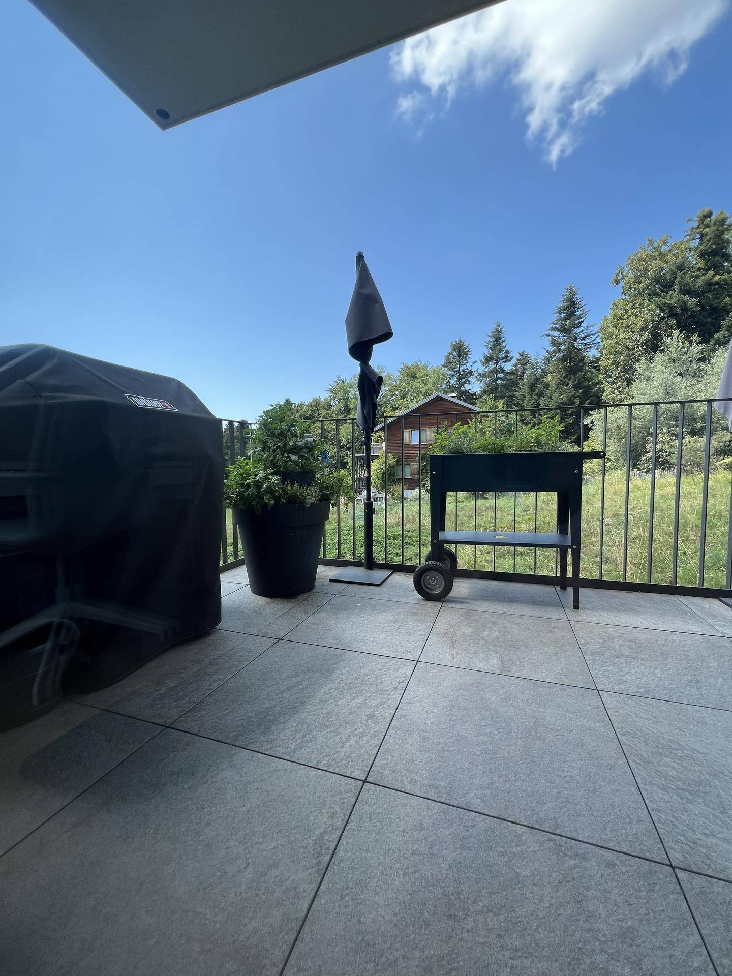 Geräumiger Balkon mit Fliesenboden, Grill und Sonnenschirm, umgeben von viel Grün.
