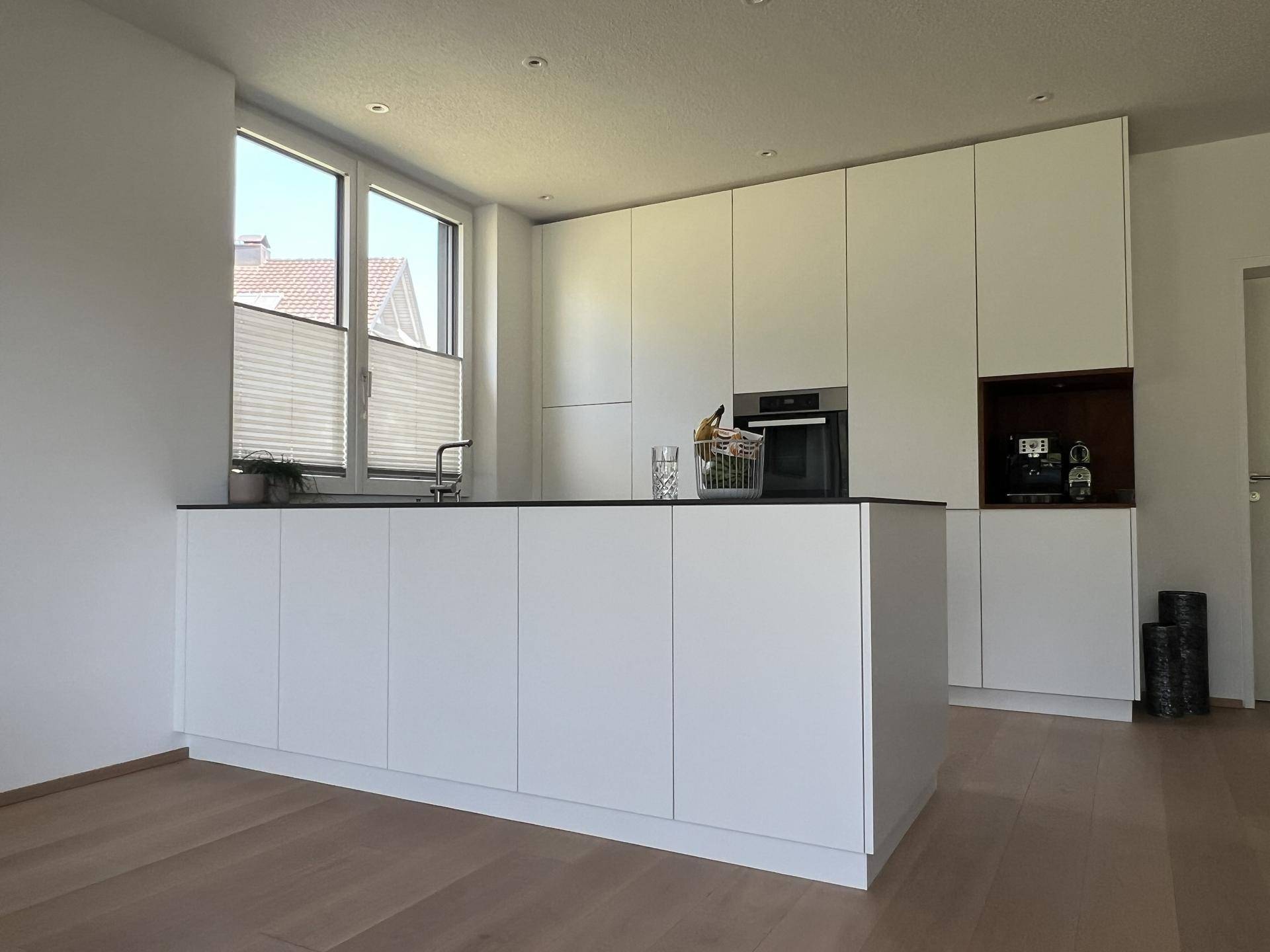 Moderne weiße Einbauküche mit Kochinsel, integriertem Backofen und Fenster über der Spüle.