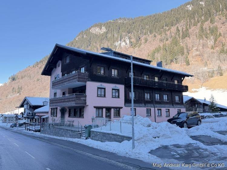 Gebäude mit rosa Fassade und Holzbalkonen, umgeben von schneebedeckten Bergen und einem Parkplatz mit Schnee.