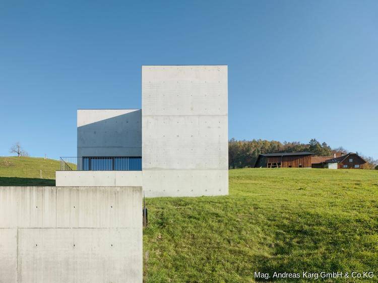 Minimalistisches Betongebäude in einer ländlichen Umgebung mit grünen Hügeln und Bäumen.