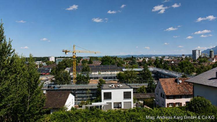 Weitläufiger Blick über eine Stadt mit grünen Baumwipfeln, Baustellen und Bergen am Horizont.