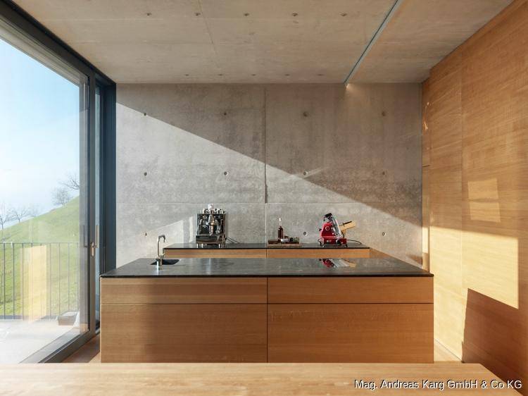 Moderne Küche mit Betonwänden, Holzelementen und großem Fenster mit Blick ins Grüne.