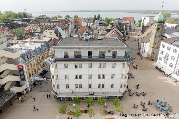 Luftaufnahme eines belebten Stadtplatzes mit Gebäuden, Kirchturm und See im Hintergrund.