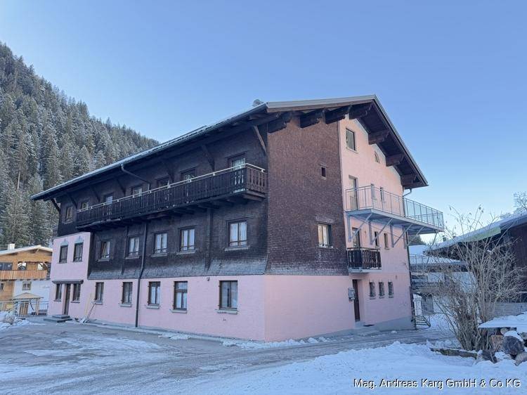 Rückseite des Gebäudes mit rosa Fassade, Holzbalkonen und einem Metallbalkon, umgeben von Bergen und Schnee.