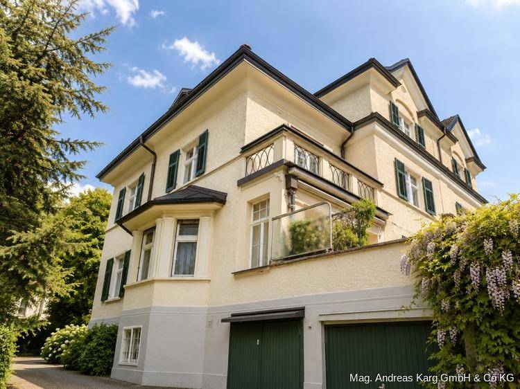 Seitenansicht der Villa mit Garagenzugang, üppiger Bepflanzung und sonnigem Himmel.