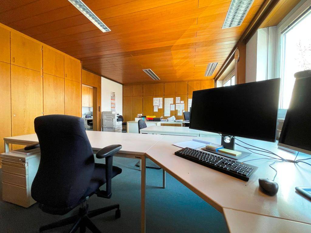 Helles Büro mit Holzdecke, Teppichboden und mehreren Arbeitsplätzen, ausgestattet mit Computern und Büromöbeln.
