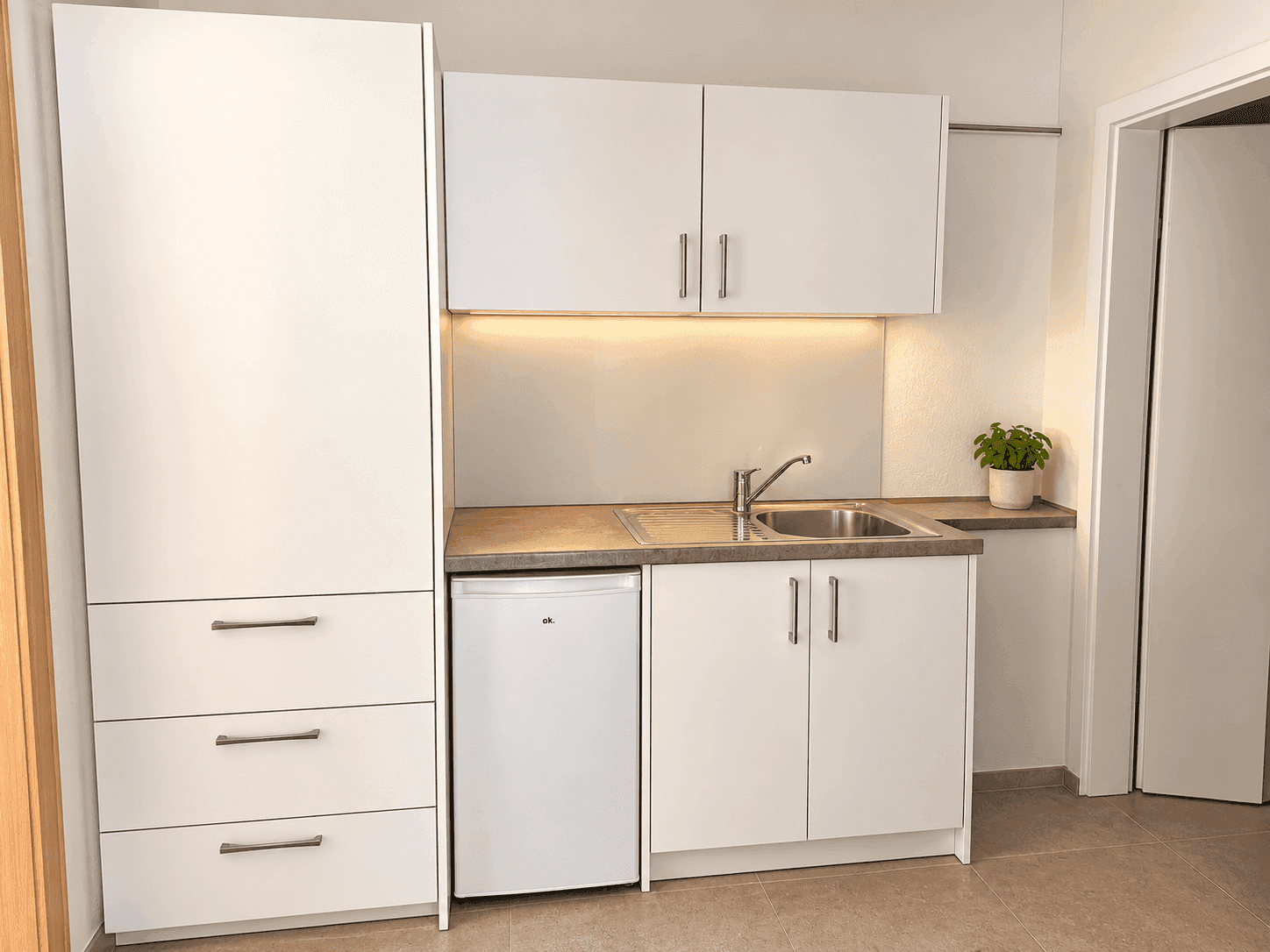 Kompakte Teeküche mit weißen Schränken, Spüle und integriertem Kühlschrank.