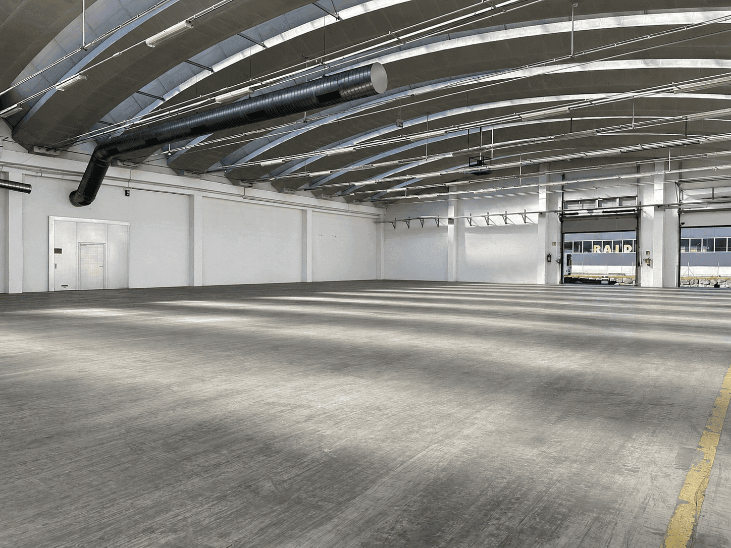 Weitläufige Industriehalle mit Betonboden, hohen Decken und einem großen Rolltor am Ende des Raumes.